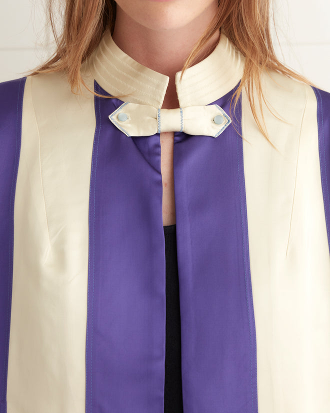 A woman wearing a Regency Jacket with satin stripes of purple and white stands confidently in black pants, adding a touch of vintage reproduction style to the tiled room.