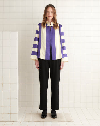 A woman wearing a Regency Jacket with satin stripes of purple and white stands confidently in black pants, adding a touch of vintage reproduction style to the tiled room.