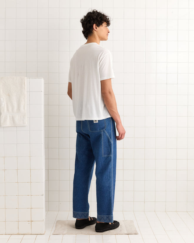 A man stands on a mat in a white tiled room, wearing the Regionals Tee, blue jeans, and black shoes.