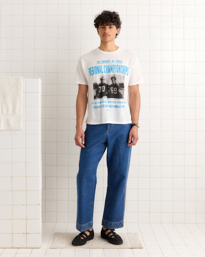 A person stands in a white tiled room, wearing the Regionals Tee styled with blue jeans and black sandals.