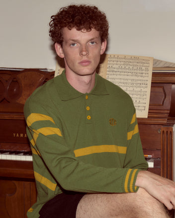 A young man with curly hair sits by a piano, wearing the Regis Polo, a green and yellow striped virgin wool sweater.