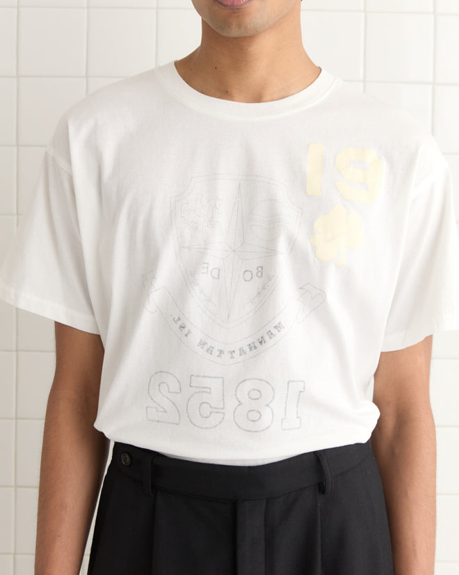 The individual is wearing a Reversible Crest Tee, a 90s-inspired shirt made from lightweight cotton, and black pants, standing against a tiled wall.