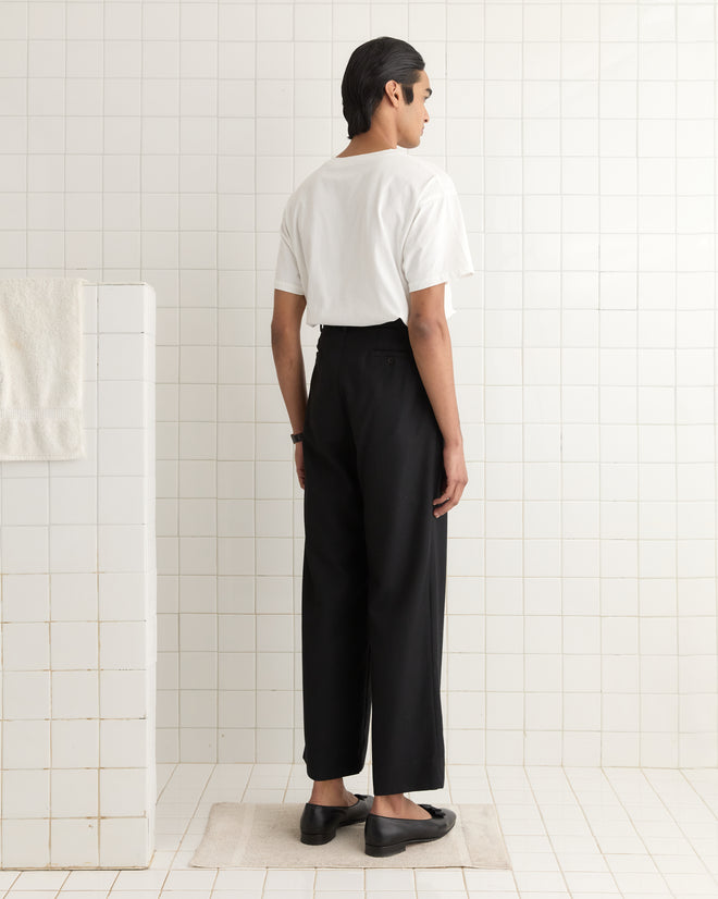 A man in a 1990s-inspired Reversible Crest Tee and black pants stands on a beige mat in a tiled room, facing away.