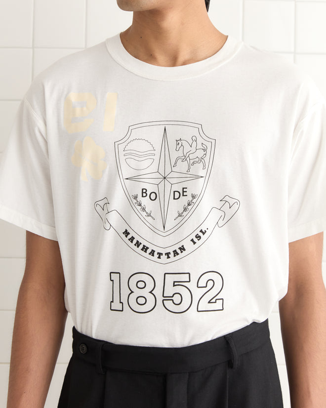 A man wears the Reversible Crest Tee, a lightweight cotton T-shirt with "BODE," a crest design, "Manhattan Isl.," and "1852," styled in a 1990s vibe.