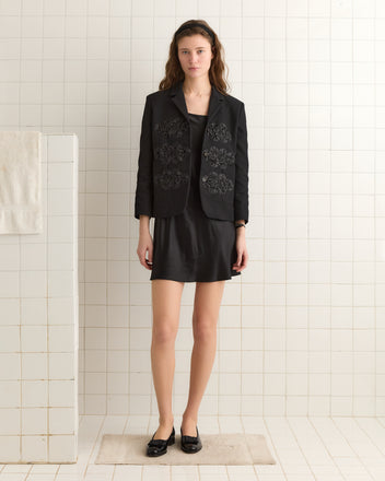 Woman wearing the Ribbon Garland Jacket - Black stands on a bath mat in a white tiled room.
