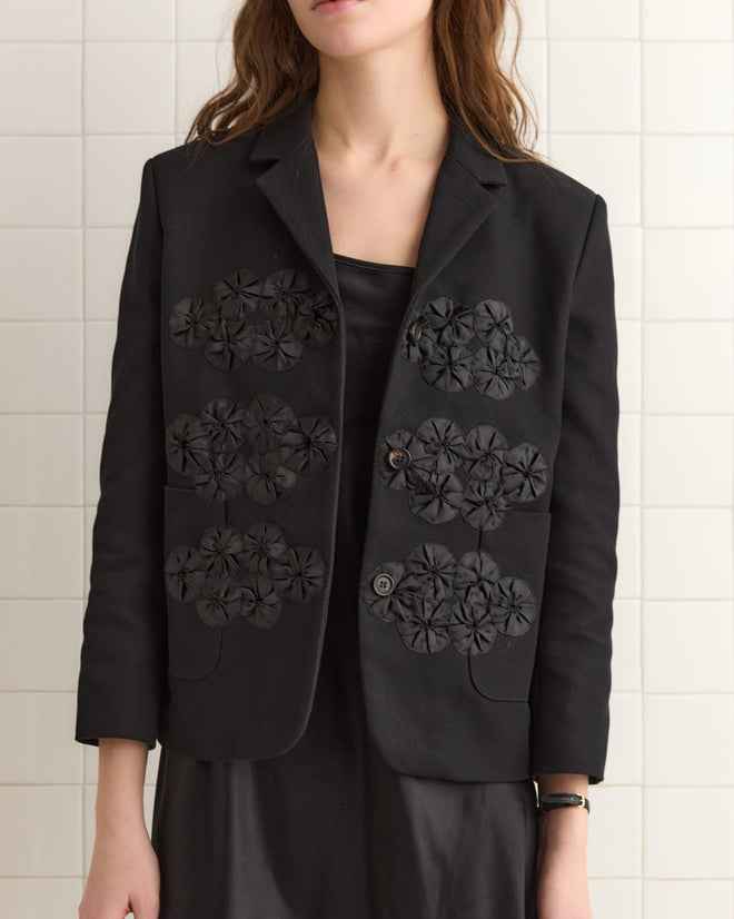 Woman wearing the Ribbon Garland Jacket - Black, a heavyweight twill women's jacket with floral appliqués, layered over a black dress and standing against a tiled wall.