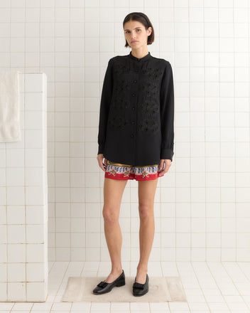 A person stands in a bathroom wearing the Ribbon Garland Silas Shirt - Black, complemented by red patterned shorts and black loafers. The outfit exudes elegance with its 100% silk fabric accentuating the ensemble.