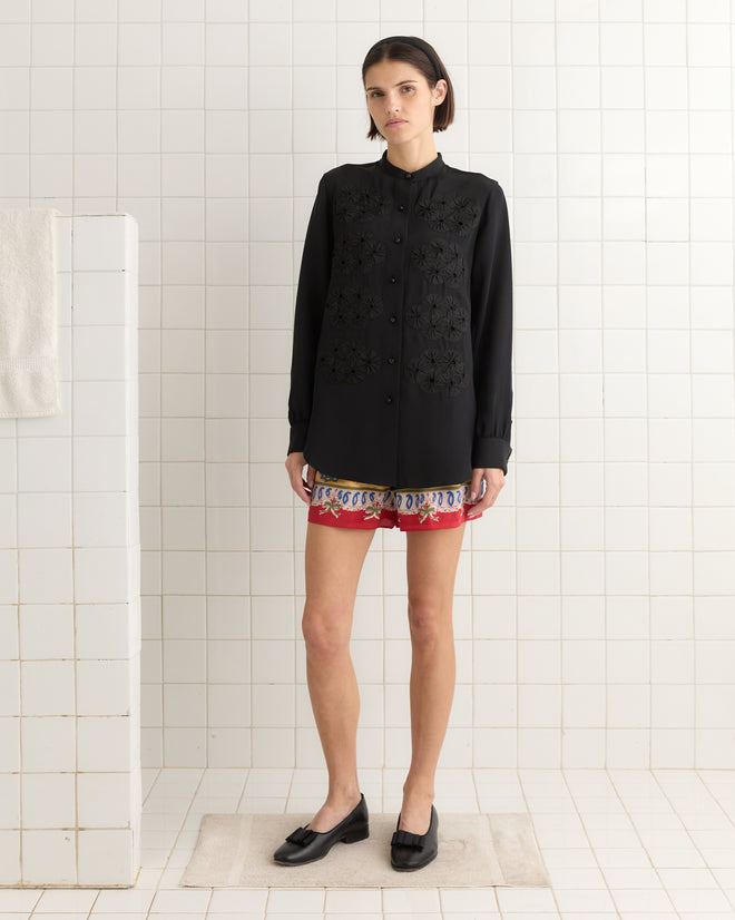 A person stands in a bathroom wearing the Ribbon Garland Silas Shirt - Black, complemented by red patterned shorts and black loafers. The outfit exudes elegance with its 100% silk fabric accentuating the ensemble.