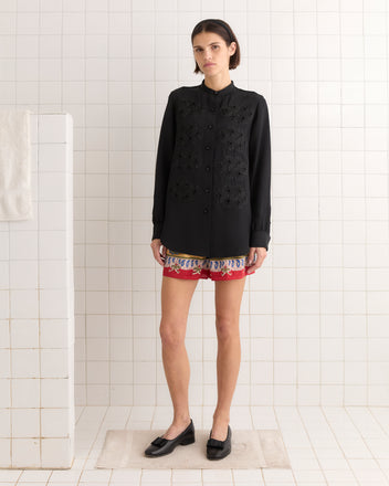 A person in a Ribbon Garland Silas Shirt - Black and red shorts stands in a tiled room, with a towel hanging on a nearby wall.