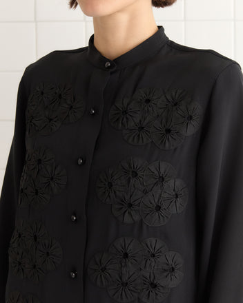 A woman in a Ribbon Garland Silas Shirt - Black, featuring textured circular patterns reminiscent of a jockey jacket, stands against a tiled wall.