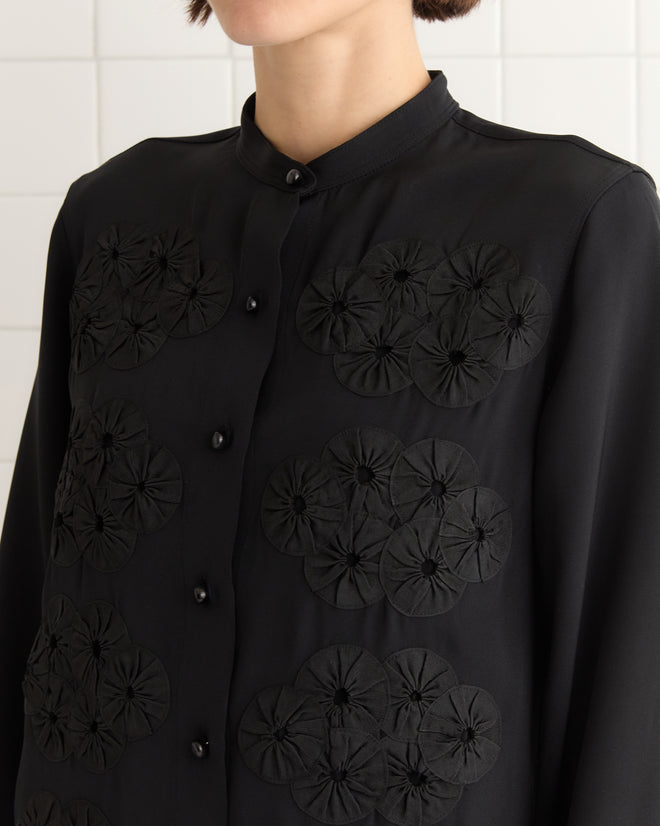 A woman in a Ribbon Garland Silas Shirt - Black, featuring textured circular patterns reminiscent of a jockey jacket, stands against a tiled wall.