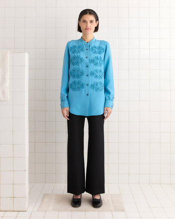 A person in a Ribbon Garland Silas Shirt - Blue with intricate ribbon appliqués and black pants stands in a tiled room, facing forward.