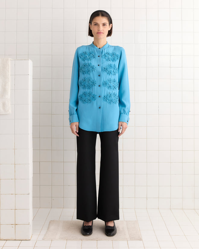 A person in a Ribbon Garland Silas Shirt - Blue with intricate ribbon appliqués and black pants stands in a tiled room, facing forward.