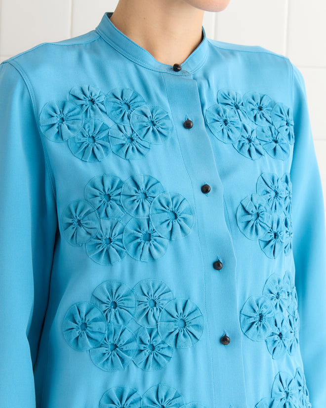 The person is wearing the Ribbon Garland Silas Shirt in blue, featuring decorative floral patterns and black buttons down the front.