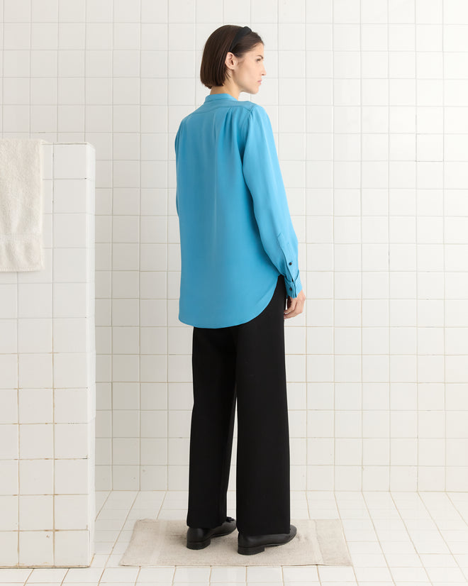 A person in a Ribbon Garland Silas Shirt - Blue and black pants stands on a light rug in a tiled room, facing left.
