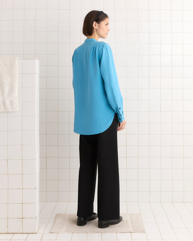 A person in a Ribbon Garland Silas Shirt - Blue and black pants stands on a bath mat in a tiled room, facing away.