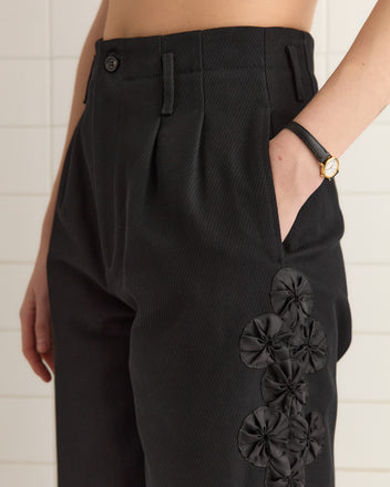 Person wearing Ribbon Garland Trousers in black, featuring floral appliqué and a wide-leg silhouette, with hand in pocket and a black watch on their wrist.