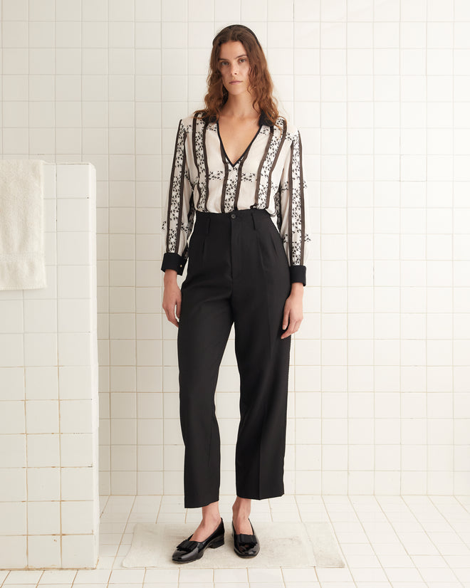 A woman stands in a tiled room wearing a black and white patterned blouse, Ribbon Murray Trousers, and black shoes.