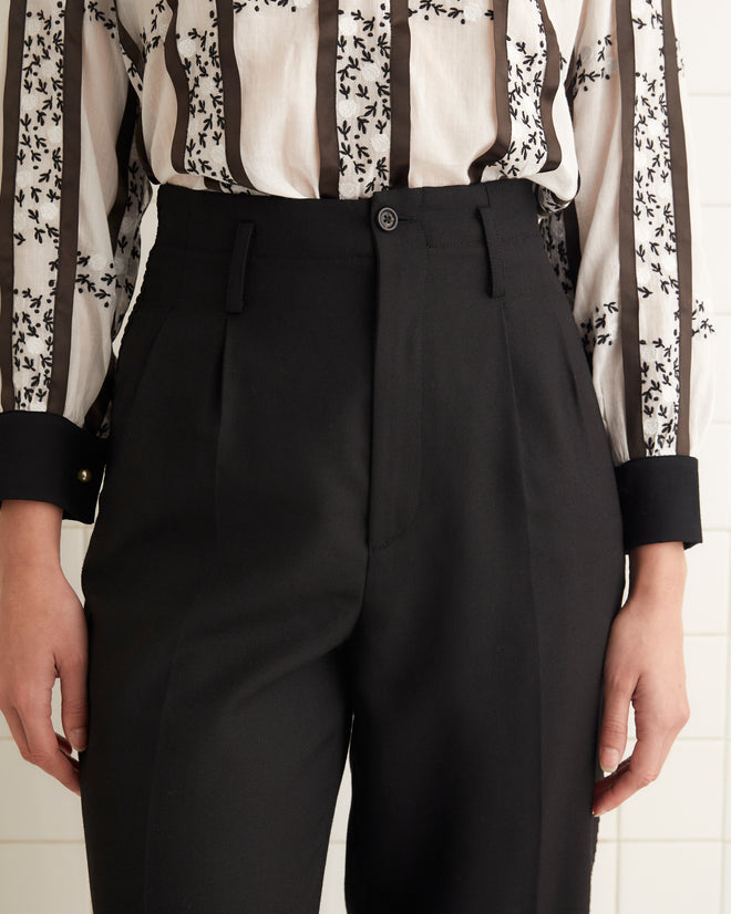 A woman stands in a tiled room wearing a black and white patterned blouse, Ribbon Murray Trousers, and black shoes.