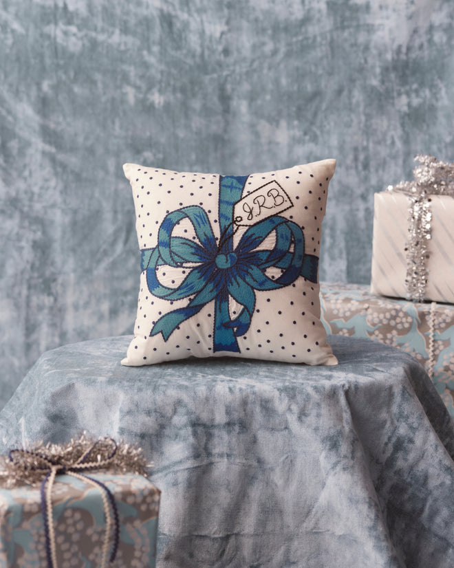 The Ribbon Pillow - Blue, featuring white embroidery with a blue ribbon design set against a textured blue backdrop and surrounded by wrapped gifts, is an elegant addition to your home goods collection.