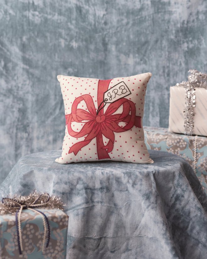 The Ribbon Pillow - Pink, featuring intricate embroidery and a charming red gift bow design, elegantly rests on a blue draped surface amidst wrapped presents—a perfect personalized gift for anyone who appreciates unique home goods.