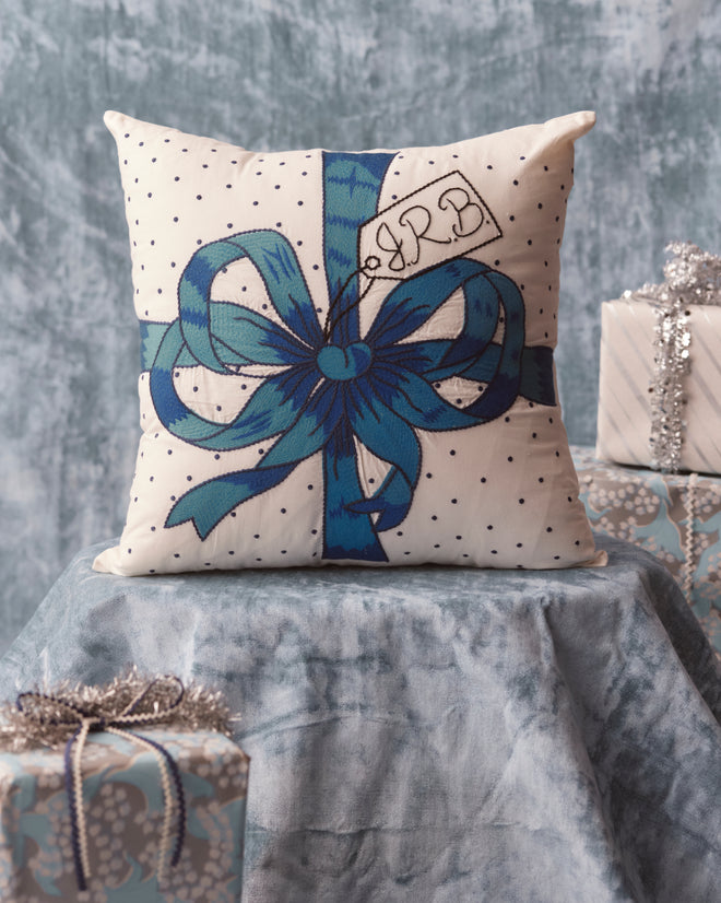 The Ribbon Pillow - Blue, featuring white embroidery with a blue ribbon design set against a textured blue backdrop and surrounded by wrapped gifts, is an elegant addition to your home goods collection.