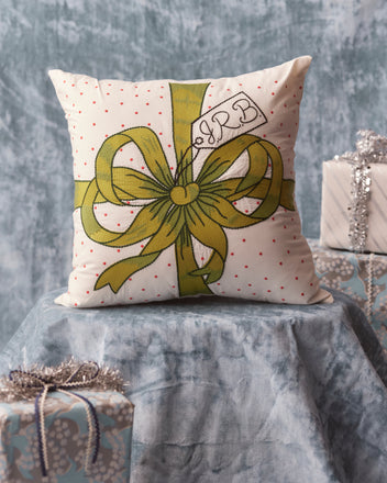 The Ribbon Pillow - Green, featuring a personalized embroidered monogram tag and a green bow design, elegantly rests on a textured blue surface, surrounded by wrapped gifts—an ideal addition to your home goods collection.