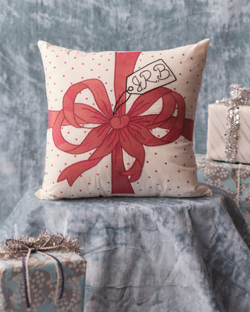 The Ribbon Pillow - Pink, featuring intricate embroidery and a charming red gift bow design, elegantly rests on a blue draped surface amidst wrapped presents—a perfect personalized gift for anyone who appreciates unique home goods.