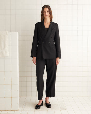 A person stands in a bathroom wearing the Gil Suit Jacket, a black double-breasted suit reminiscent of 1920s style, paired with matching pants and black shoes. The ensemble features a wide shawl collar, crafted from luxurious virgin wool. The background showcases white tiled walls and a towel hanging on a rack.