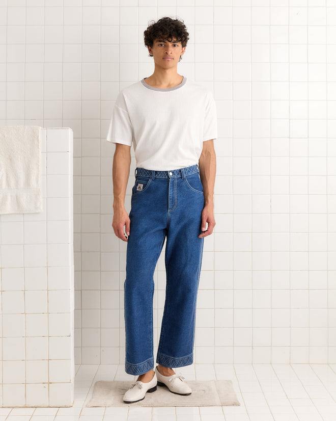 A man stands indoors against a tiled white wall wearing the Rib Boxy Tee—a vintage white shirt with a grey ringer neckline—paired with blue jeans and white shoes.