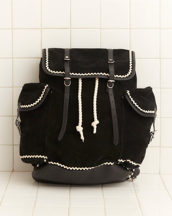 Rickrack Bookbag in black suede adorned with white rickrack trim and rope drawstrings, set against a tiled background.