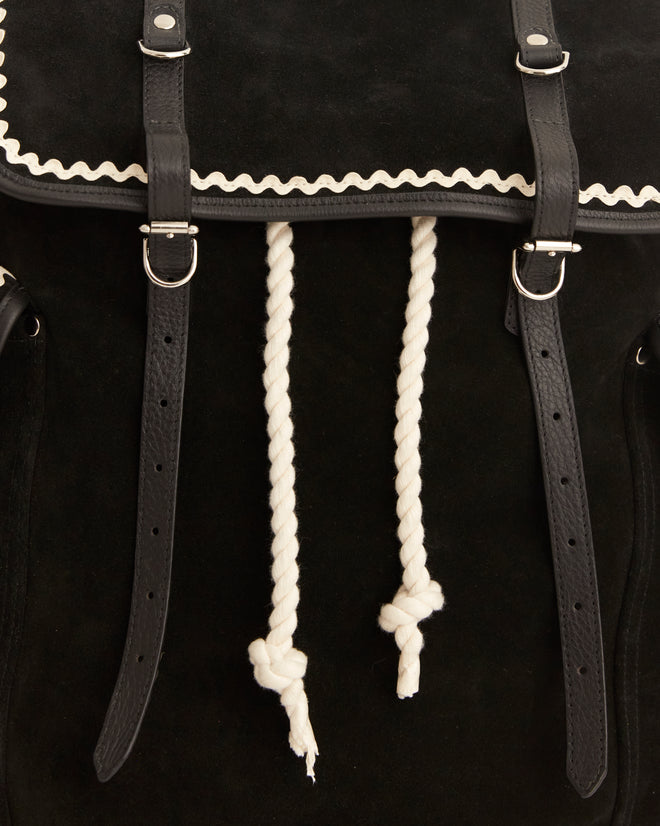 The Rickrack Bookbag, featuring black suede with white trim and rope drawstrings, is set against a tiled background, exuding the charm of a vintage rucksack.