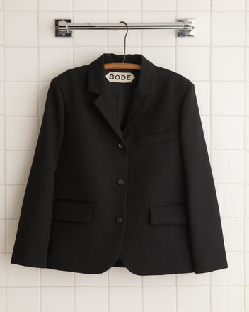 Dressed in a sophisticated Riding Twill Lillian Jacket - Black, a person stands gracefully against a patterned curtain backdrop.