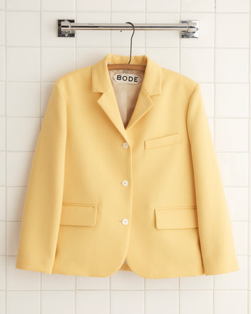 A Riding Twill Lillian Jacket in a yellow shade, adorned with white buttons, hangs elegantly on a tiled wall.