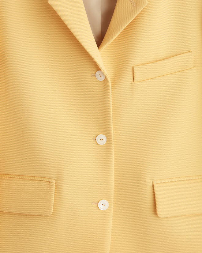 A Riding Twill Lillian Jacket in a yellow shade, adorned with white buttons, hangs elegantly on a tiled wall.