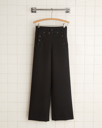 The Riding Twill Sailor Trousers in black, featuring a high-waisted wide-leg fit with button detailing and evoking the silhouette of 1930s riding breeches, hang on a wooden hanger against a tiled wall.