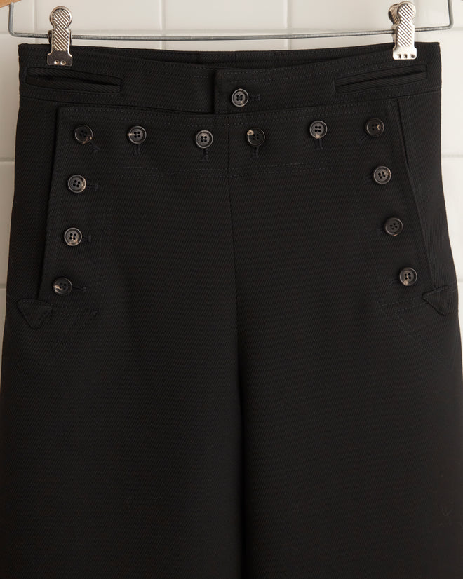 The Riding Twill Sailor Trousers in black, featuring a high-waisted wide-leg fit with button detailing and evoking the silhouette of 1930s riding breeches, hang on a wooden hanger against a tiled wall.