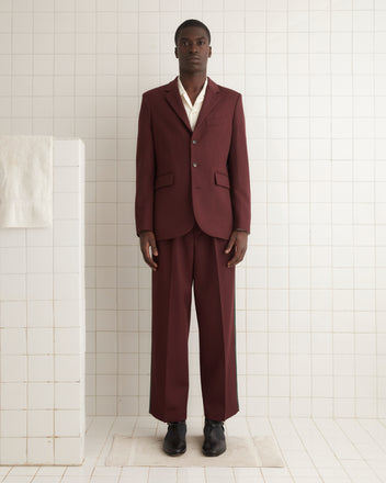 A person in a Riding Twill Suit Jacket in burgundy, featuring pearl buttons, and a crisp white shirt stands in a tiled room, where a towel hangs on the left wall.