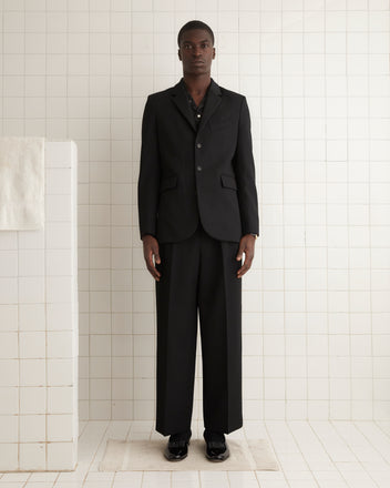 A person wearing the Riding Twill Suit Jacket in black stands in front of a patterned curtain, looking towards the camera.