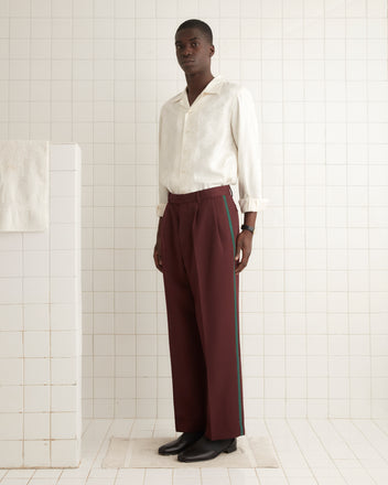 A person in a maroon suit, paired with Riding Twill Braided Trousers, sits on a wooden chair against a vintage wallpaper backdrop.