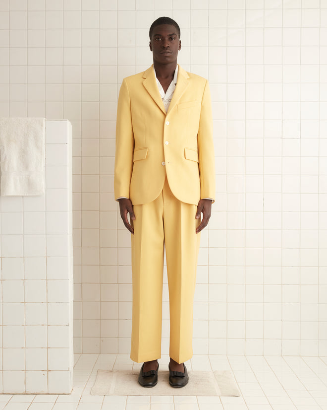 A person in a Riding Twill Suit Jacket - Yellow, paired with a white scarf, sits on a stool against a brown background adorned with dried flowers, the ensemble's pearl buttons subtly catching the light.