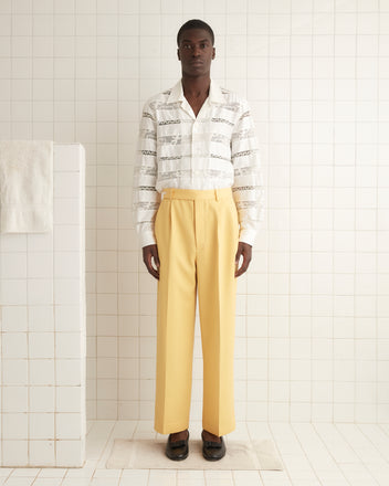 A person in a white patterned shirt with pearl buttons and Riding Twill Trousers in yellow stands against a tiled wall, exuding vintage style charm.