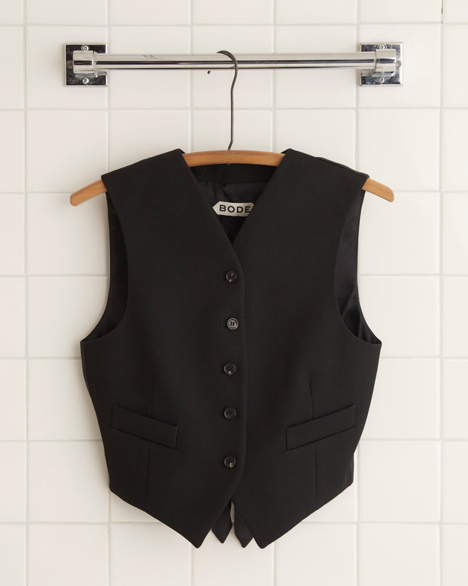 The Riding Twill Waistcoat in black hangs elegantly on a wooden hanger against a backdrop of white tiled wall, evoking the timeless charm reminiscent of 1930s riding breeches.