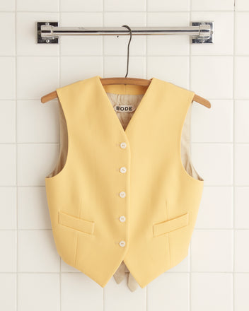 A person wearing the Riding Twill Waistcoat in yellow, with a vintage flair, holds a matching jacket while standing against a wooden backdrop.