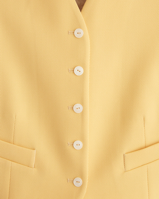 A person wearing the Riding Twill Waistcoat in yellow, with a vintage flair, holds a matching jacket while standing against a wooden backdrop.