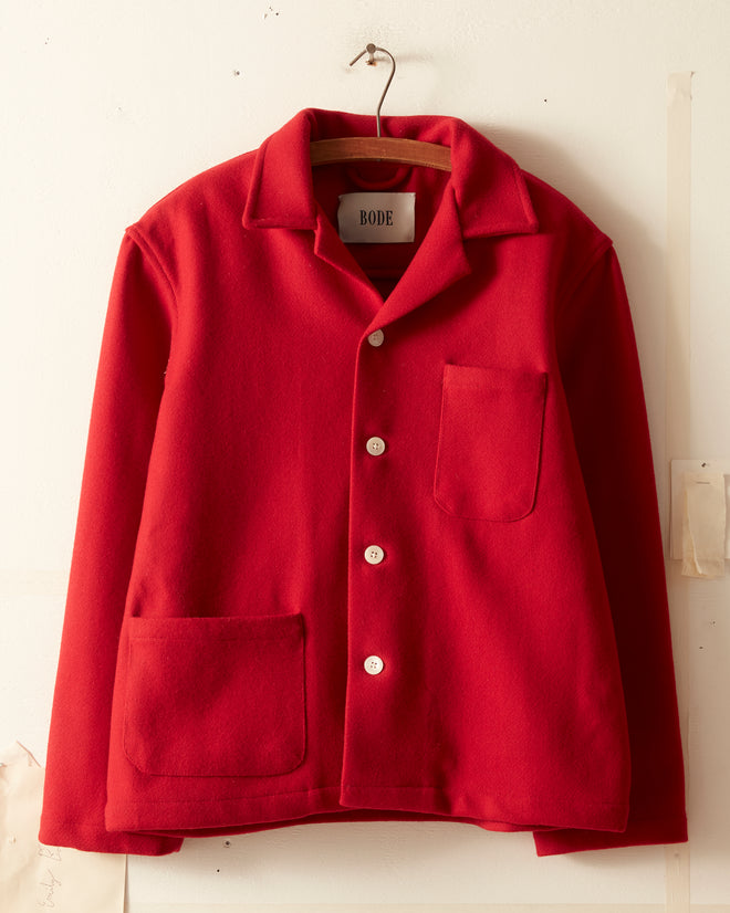 A Rioja red, long-sleeved overshirt with white buttons, two large front pockets, and a collar hangs on a hanger against a white wall. The inside of the collar features the 