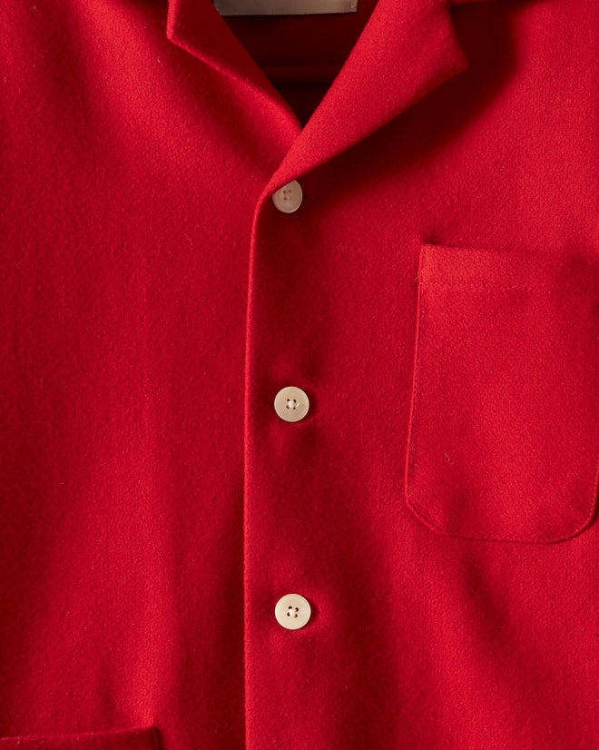 A Rioja red, long-sleeved overshirt with white buttons, two large front pockets, and a collar hangs on a hanger against a white wall. The inside of the collar features the 