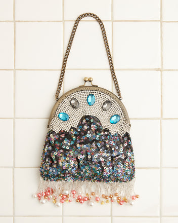 The Ritzy Bag features a chain strap and displays an eye-catching sequined design adorned with blue and clear gemstones, multicolored details, and a fringe of pearls and beads, all set against a tiled backdrop.