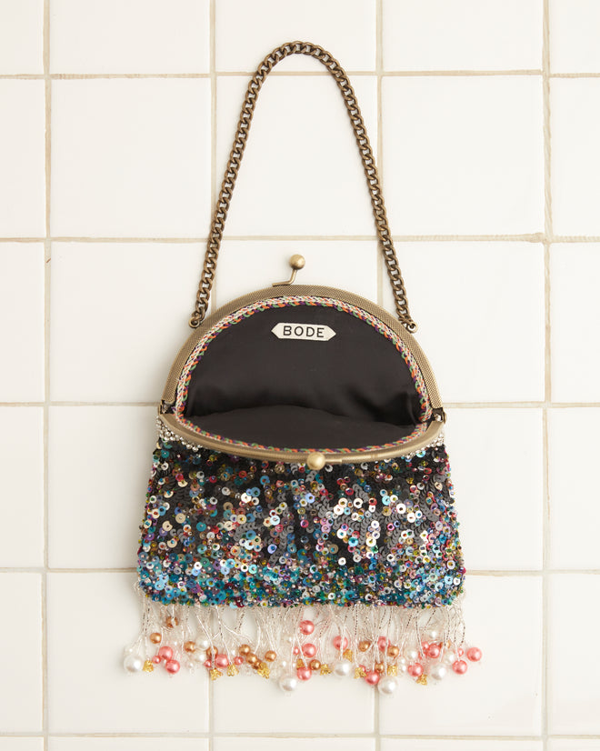 The Ritzy Bag features a chain strap and displays an eye-catching sequined design adorned with blue and clear gemstones, multicolored details, and a fringe of pearls and beads, all set against a tiled backdrop.