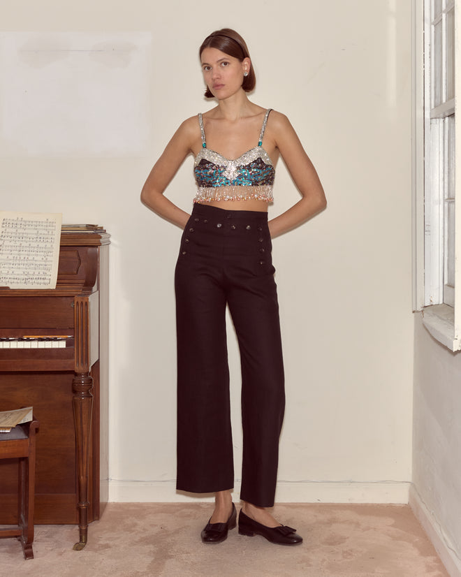 A woman wearing the Ritzy Bra and high-waisted black pants stands by a piano in a softly lit room.
